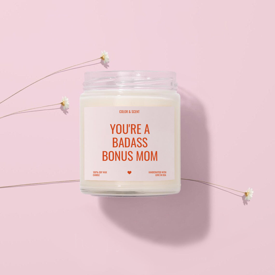 A white candle labeled "You're a Badass Bonus Mom" is placed on a pink background, surrounded by small white flowers.