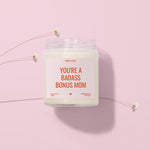 A white candle labeled "You're a Badass Bonus Mom" is placed on a pink background, surrounded by small white flowers.