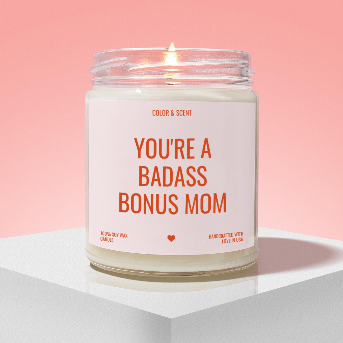A white candle with a pink background, featuring a label that reads "YOU'RE A BADASS BONUS MOM" in bold orange text.
