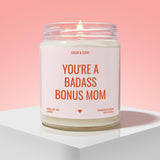 A white candle with a pink background, featuring a label that reads "YOU'RE A BADASS BONUS MOM" in bold orange text.