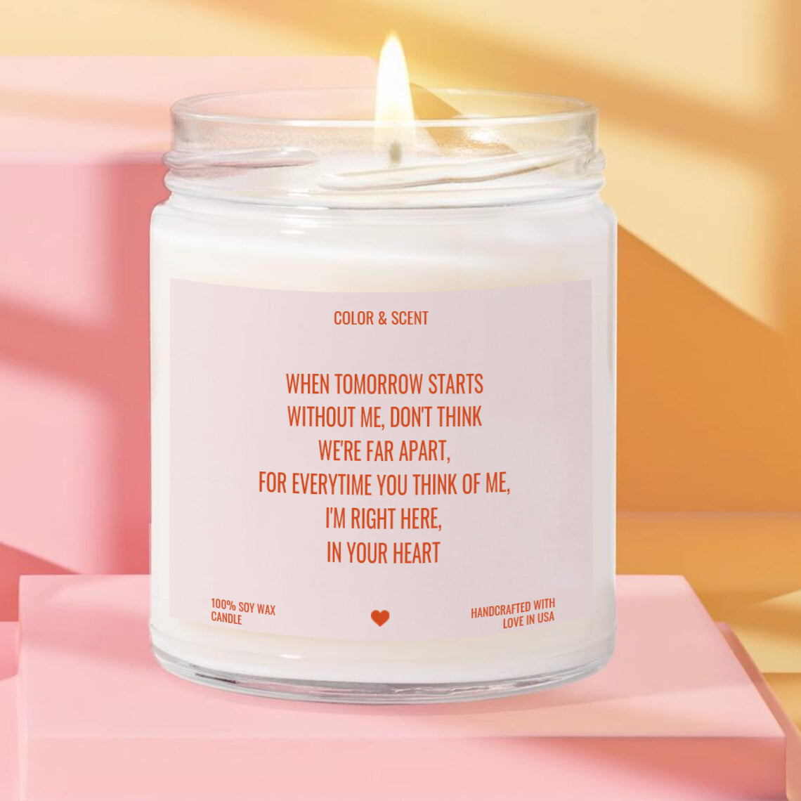 a white candle with a pink background, and it has a lit flame. The candle has a label with a message about love and distance.